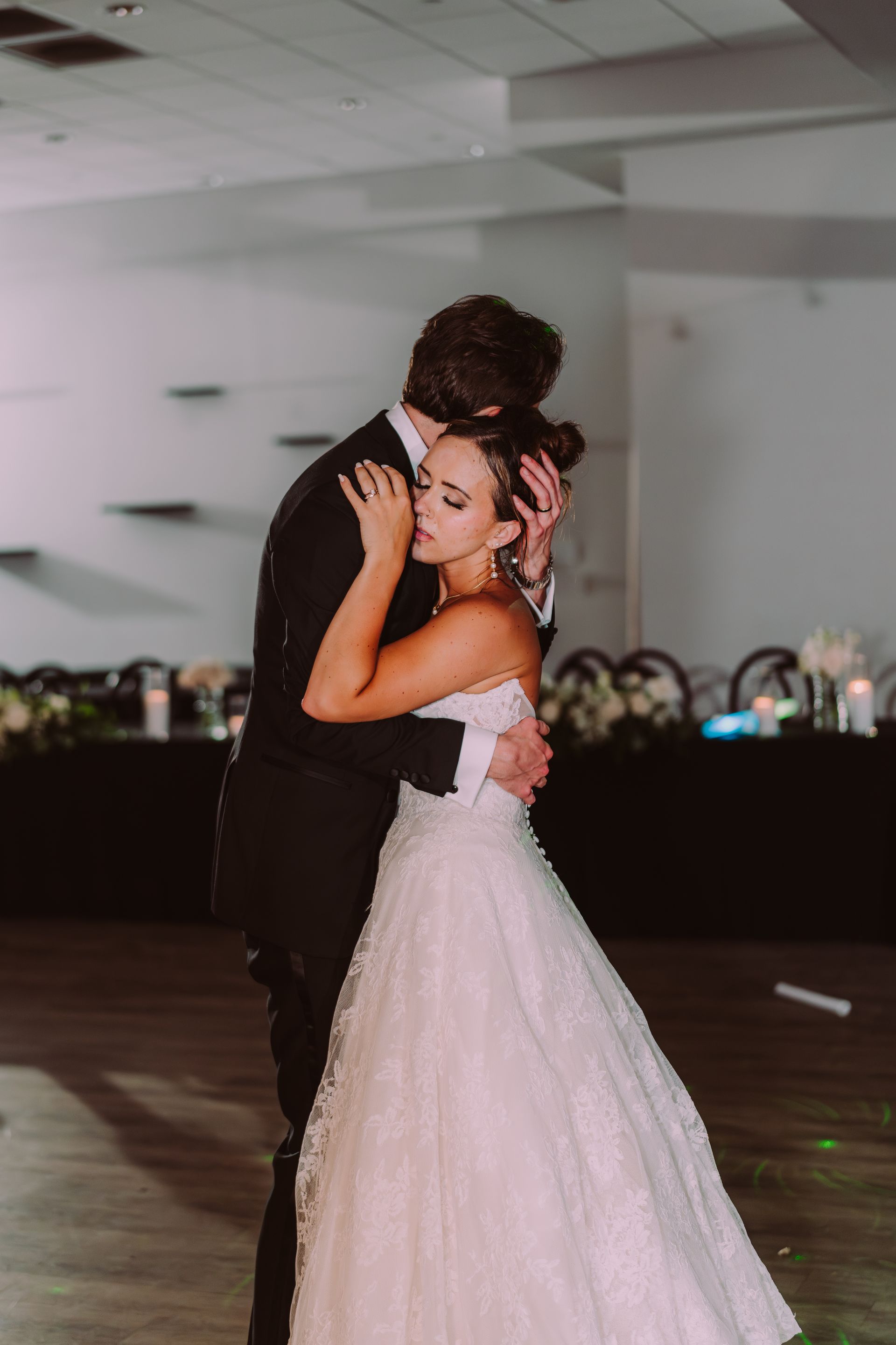 Bride and groom dance, embraced, at wedding reception. Bride wears a strapless white gown; groom, a black suit.