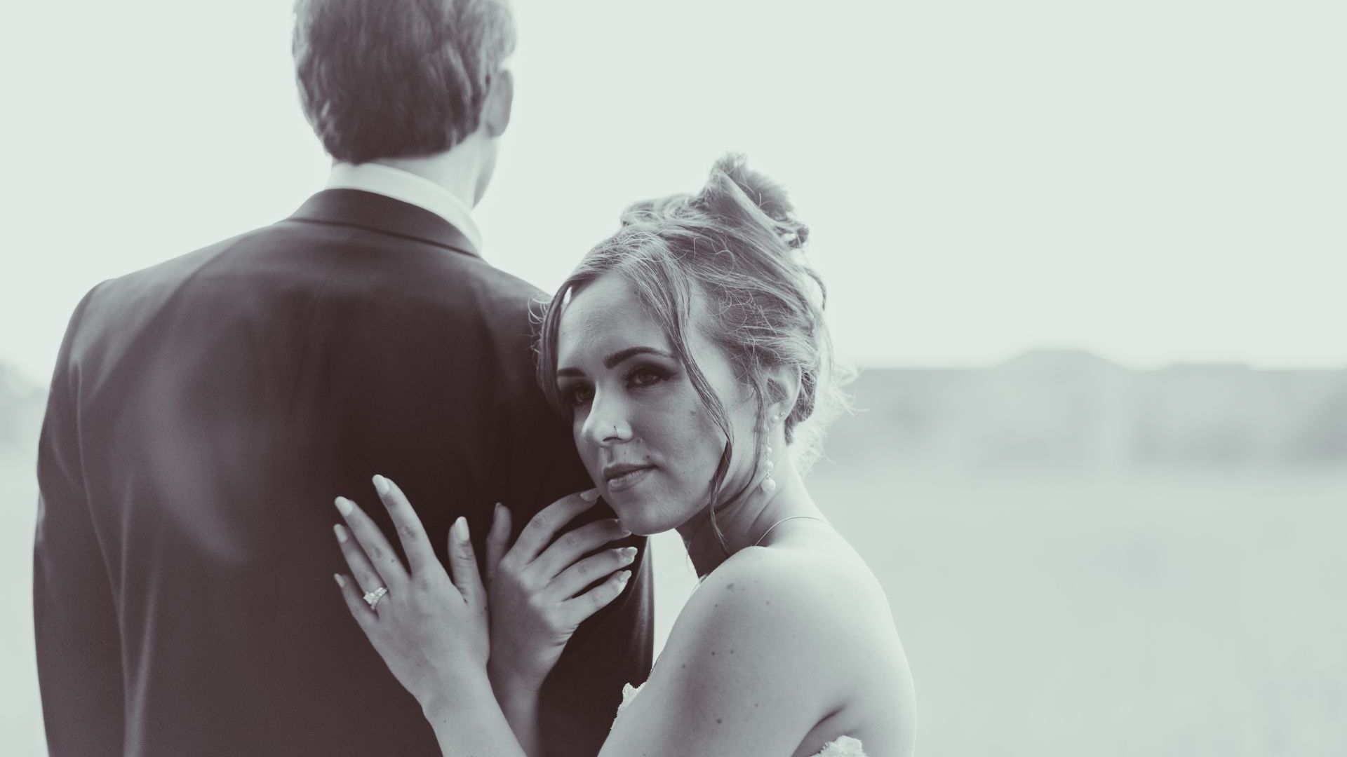 Bride embracing groom outdoors, looking back toward the camera with a soft smile