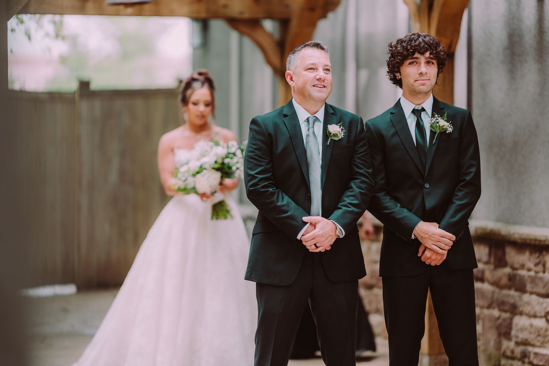 Three people standing outdoors at a wedding, with a bride in a white gown holding a bouquet behind two men in dark suits