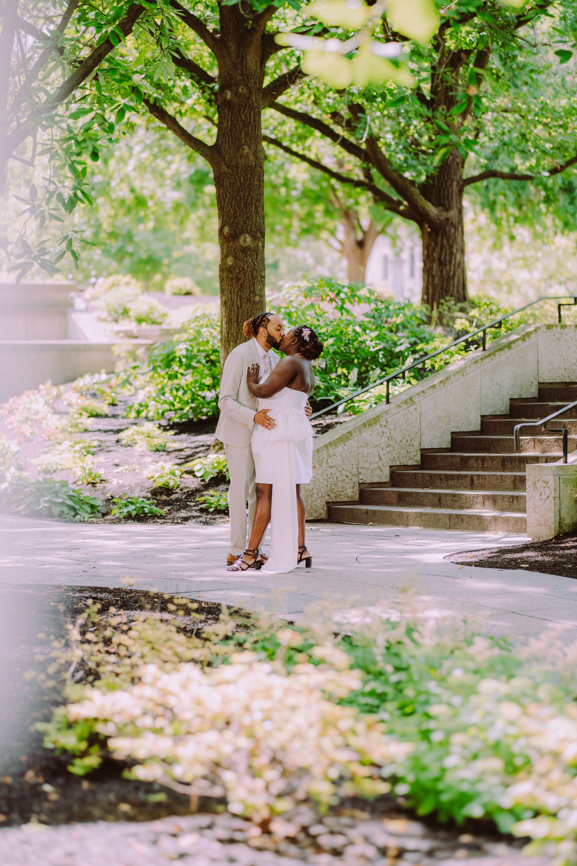 Couple embracing on a tree-lined garden path beside stone steps and greenery