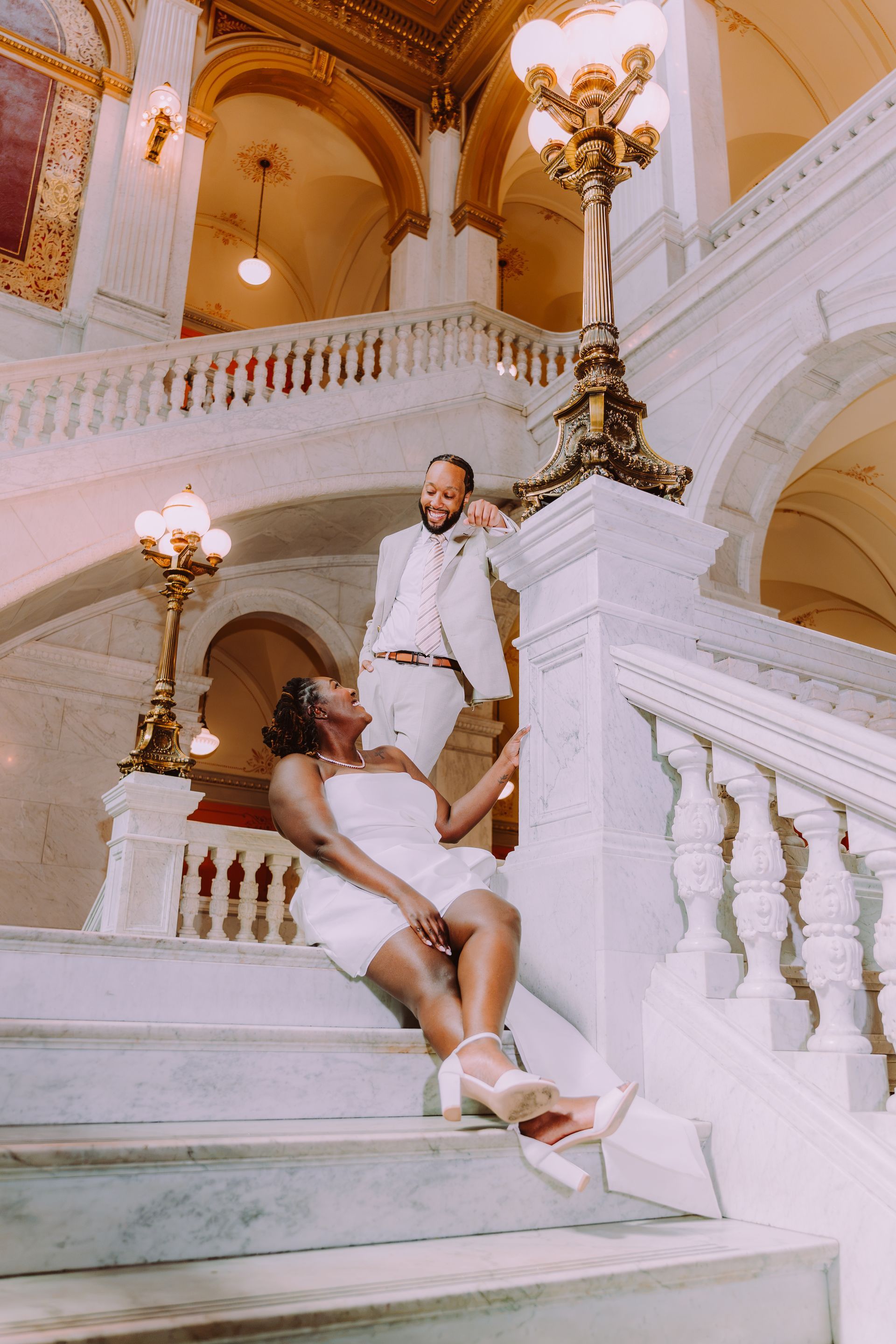 Couple in white outfits on an ornate staircase with gold chandeliers and arches