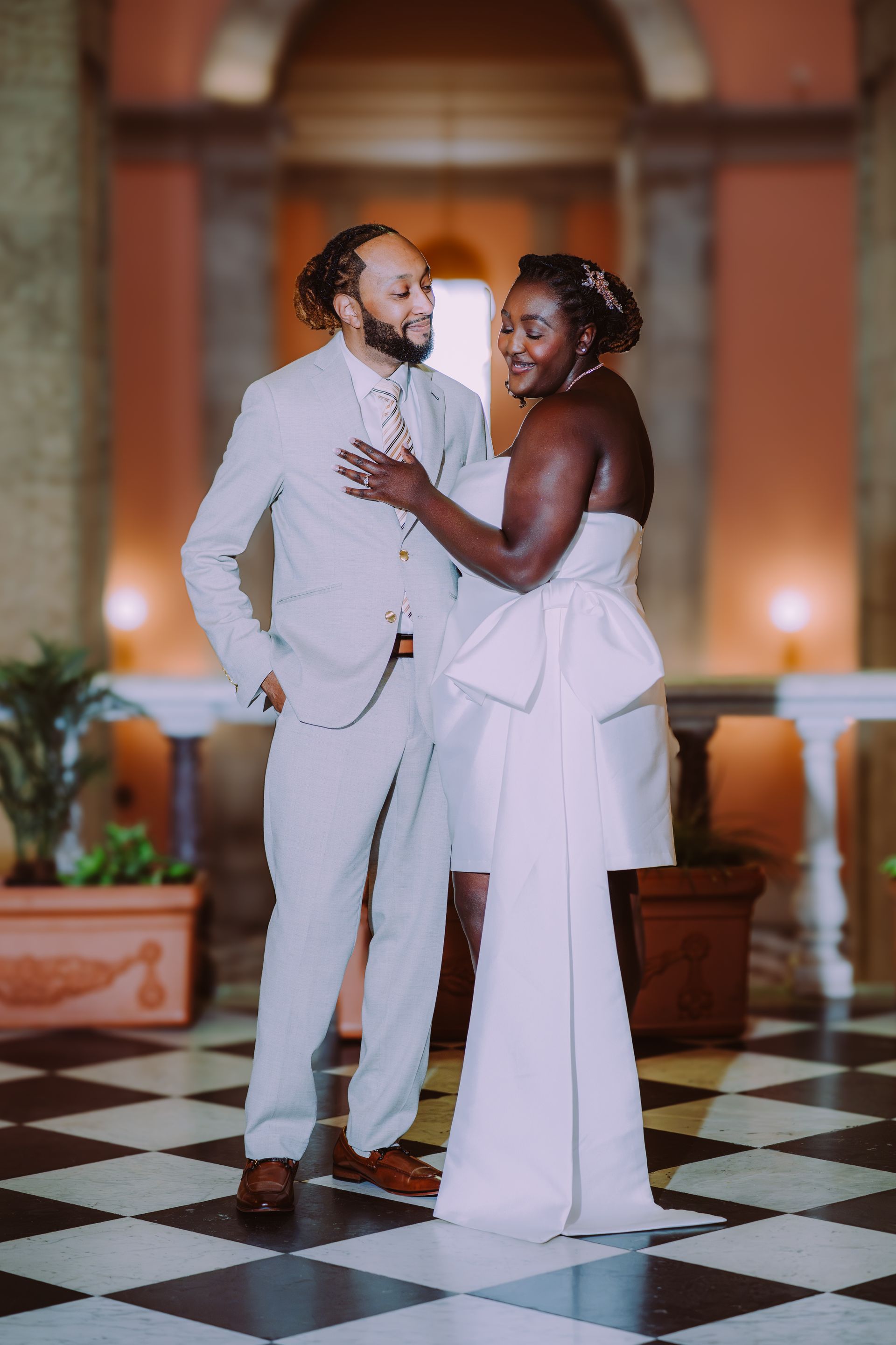 Two people in white formal wear posing together in an ornate hall with a black-and-white checkered floor.
