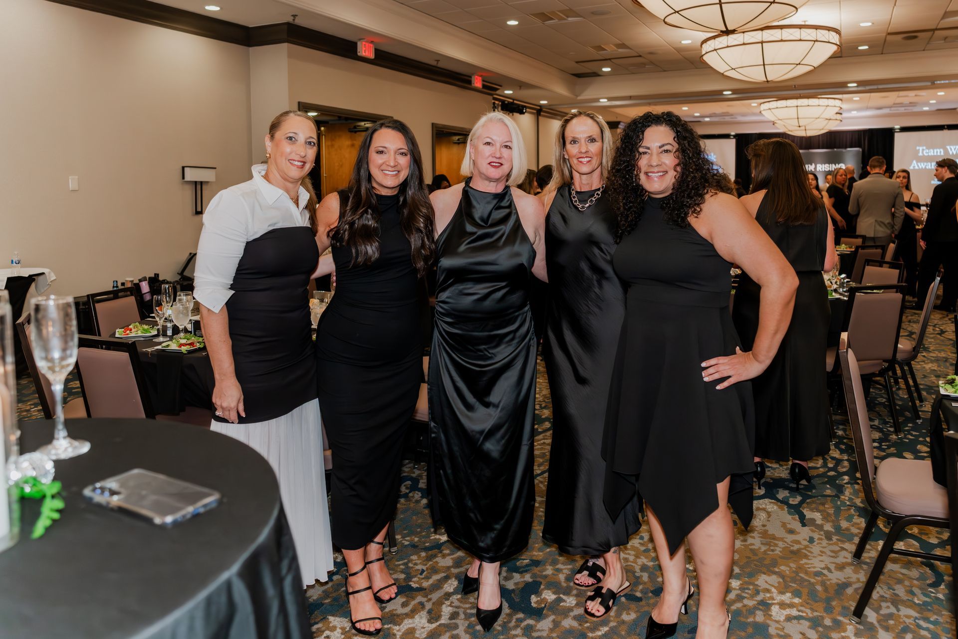 Five people stand smiling in a ballroom wearing formal attire, primarily in black, at a social or corporate event.