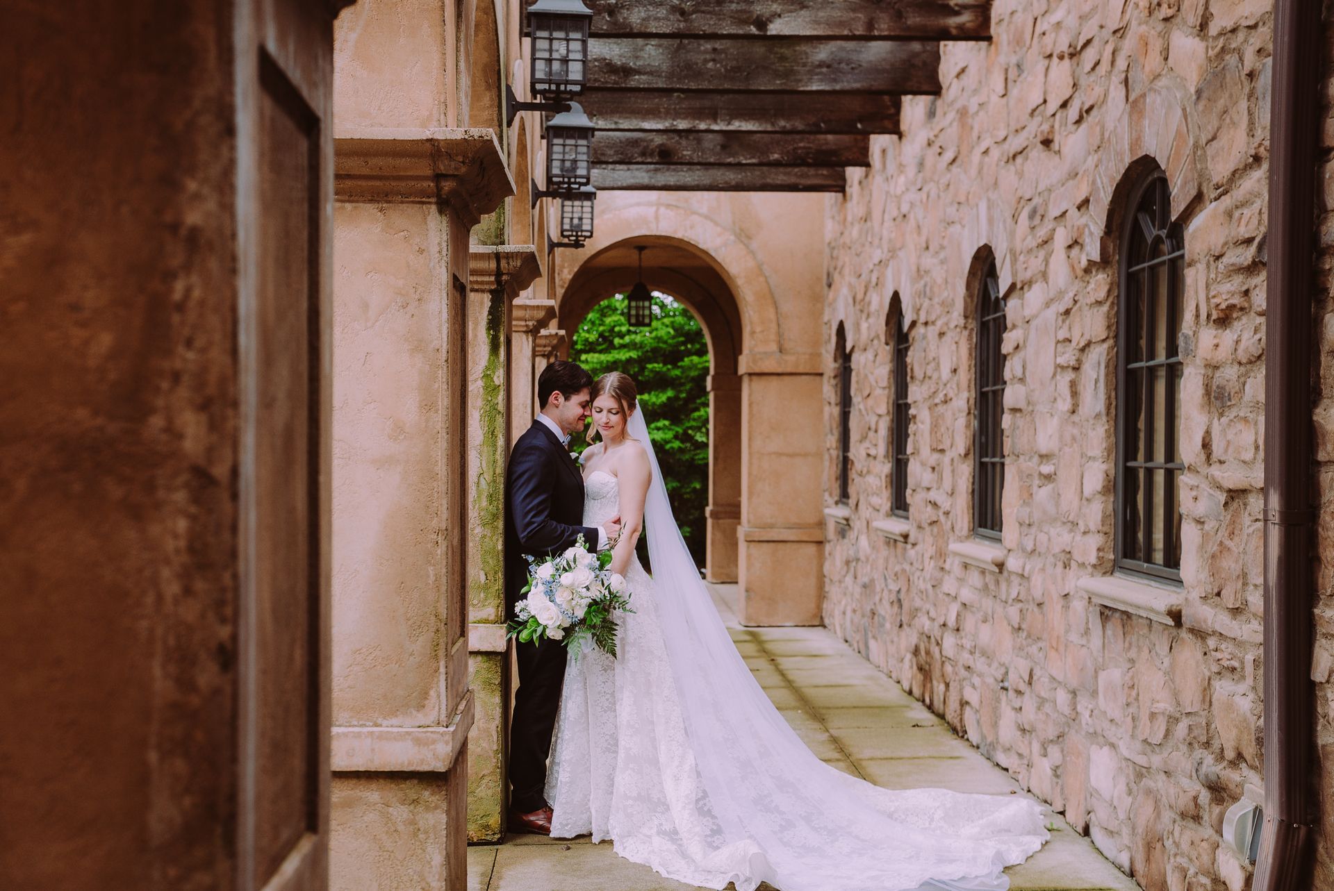 Bride and groom kiss in a stone courtyard beside an arched walkway