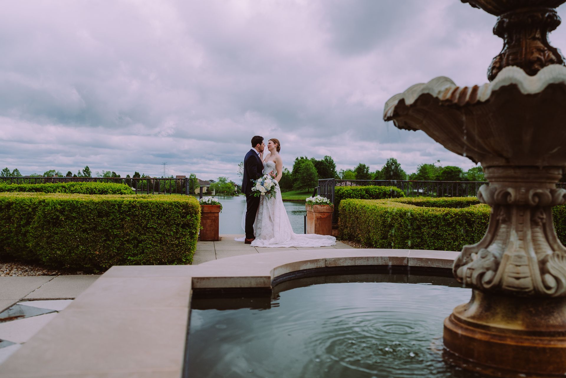 Bride and groom standing by a fountain in a garden with hedges and a cloudy sky