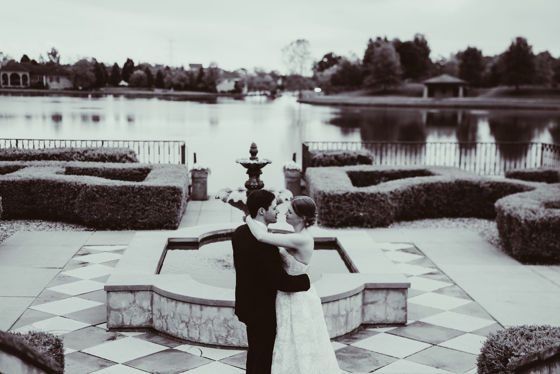 Couple dancing on a tiled terrace by a lake, with hedges and fountain in the background.