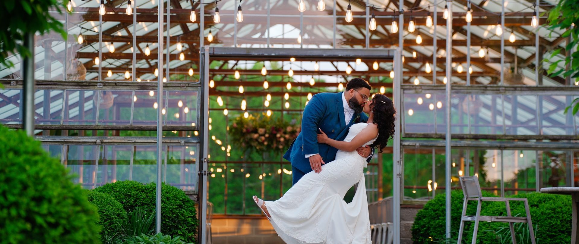 Newlyweds share a kiss in a greenhouse. The groom wears a blue suit, and the bride wears a white dress;