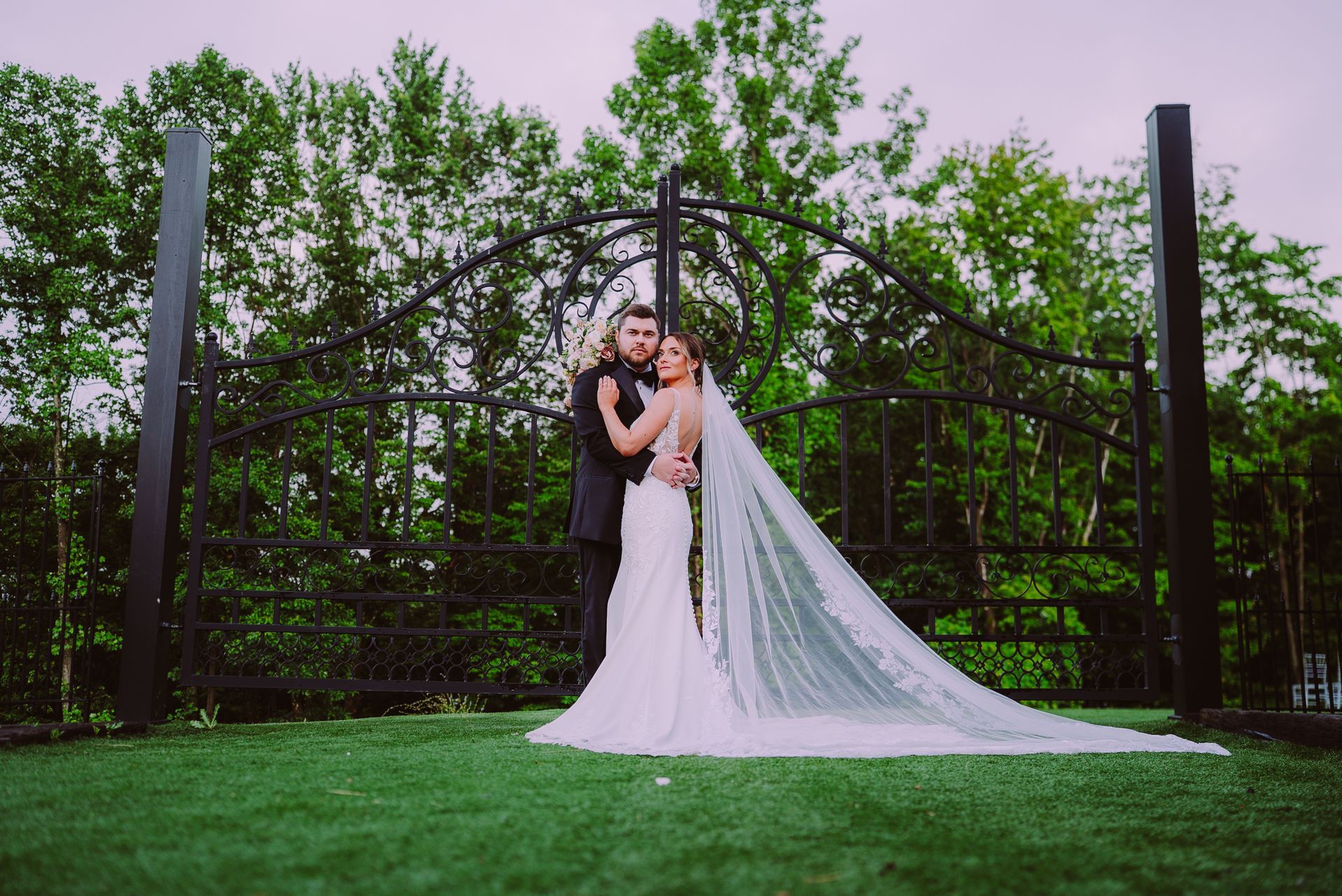 Bride and groom embracing in a garden, with a long white veil and lush green trees behind them.