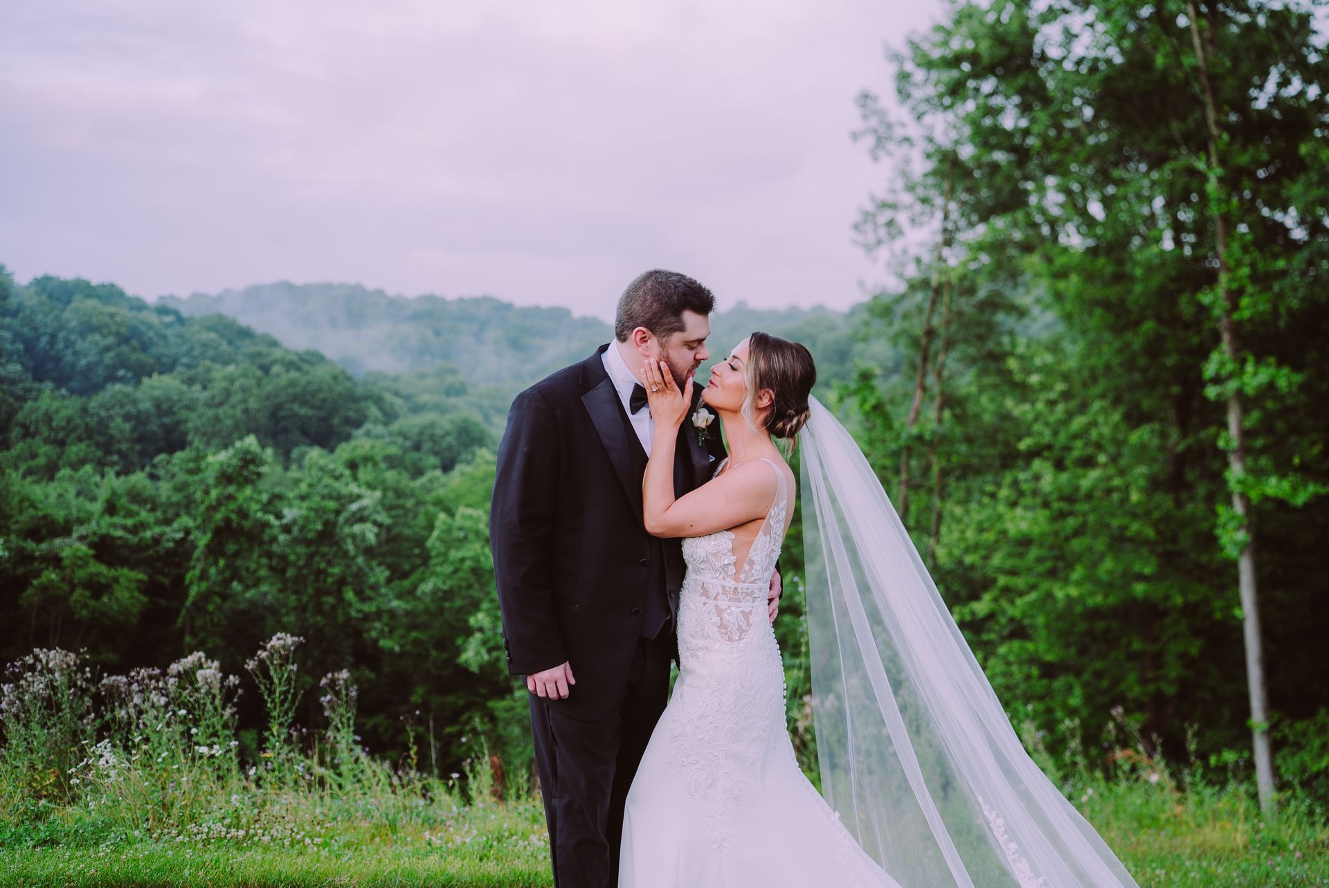 Bride and groom embrace and kiss in a lush green field, with hills and trees in the background.