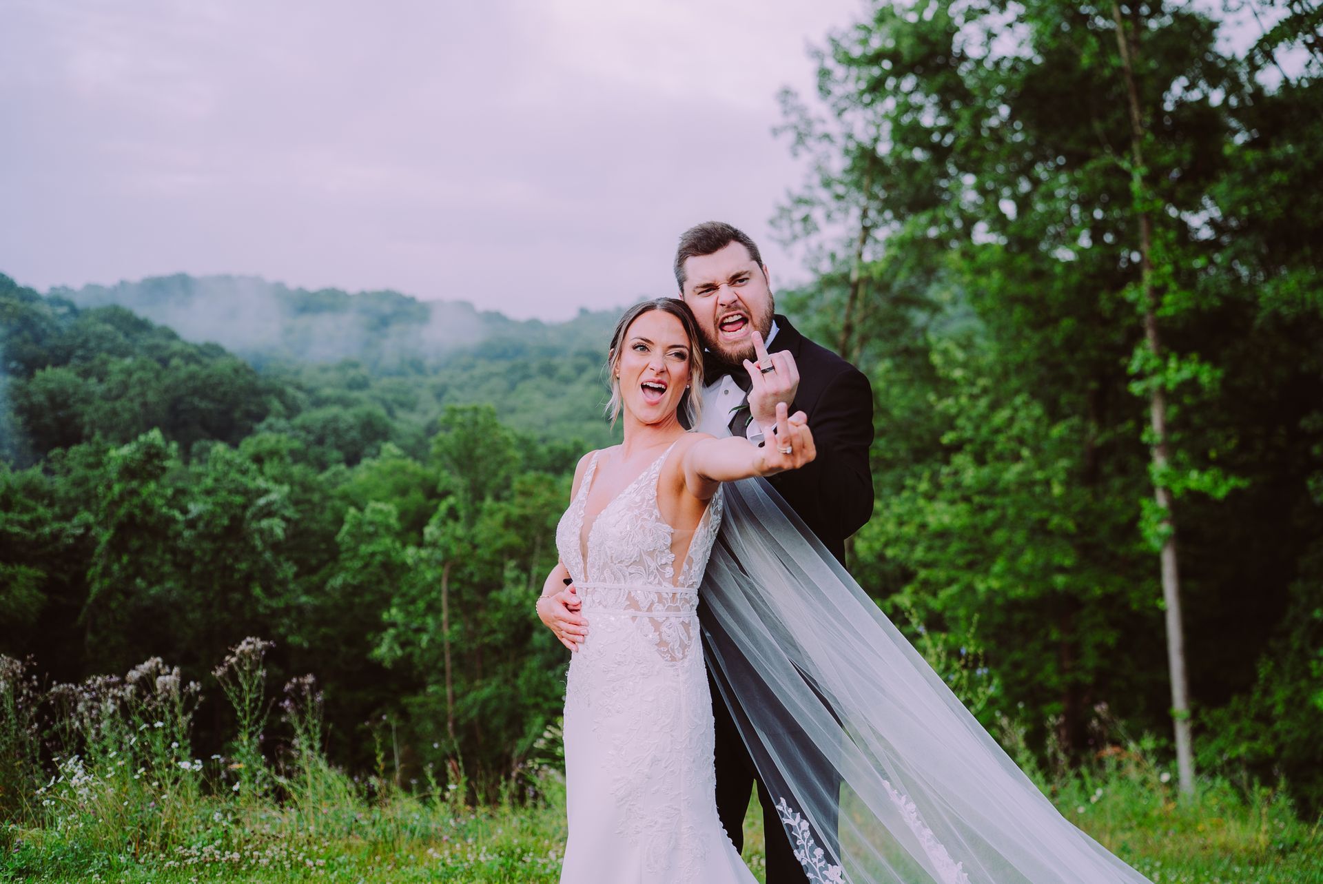Couple embracing in a white wedding dress and black suit outdoors with lush green trees and misty hills