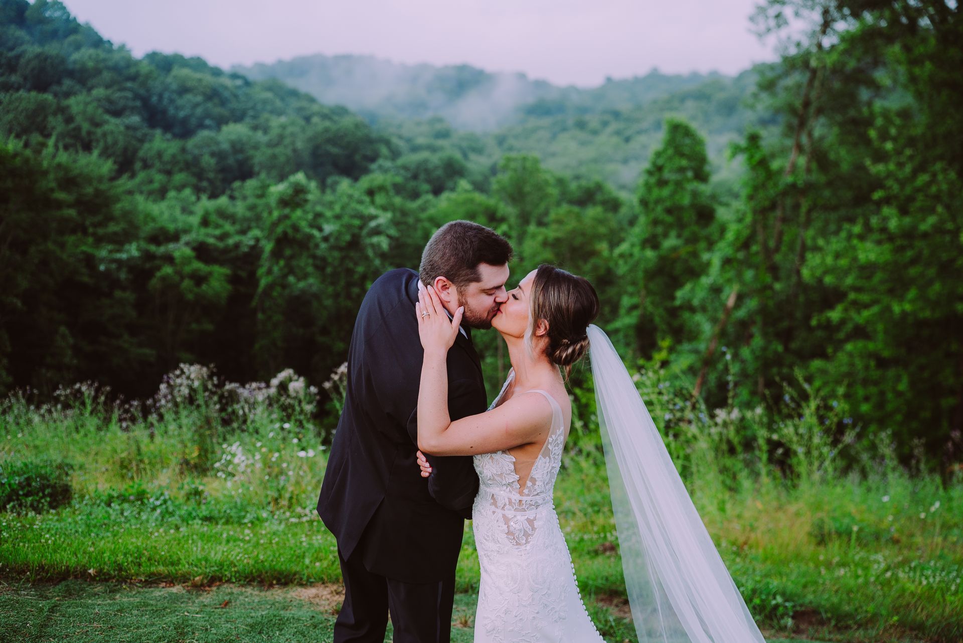 Couple kissing in a white wedding dress and dark suit, standing in a green field with misty hills behind them