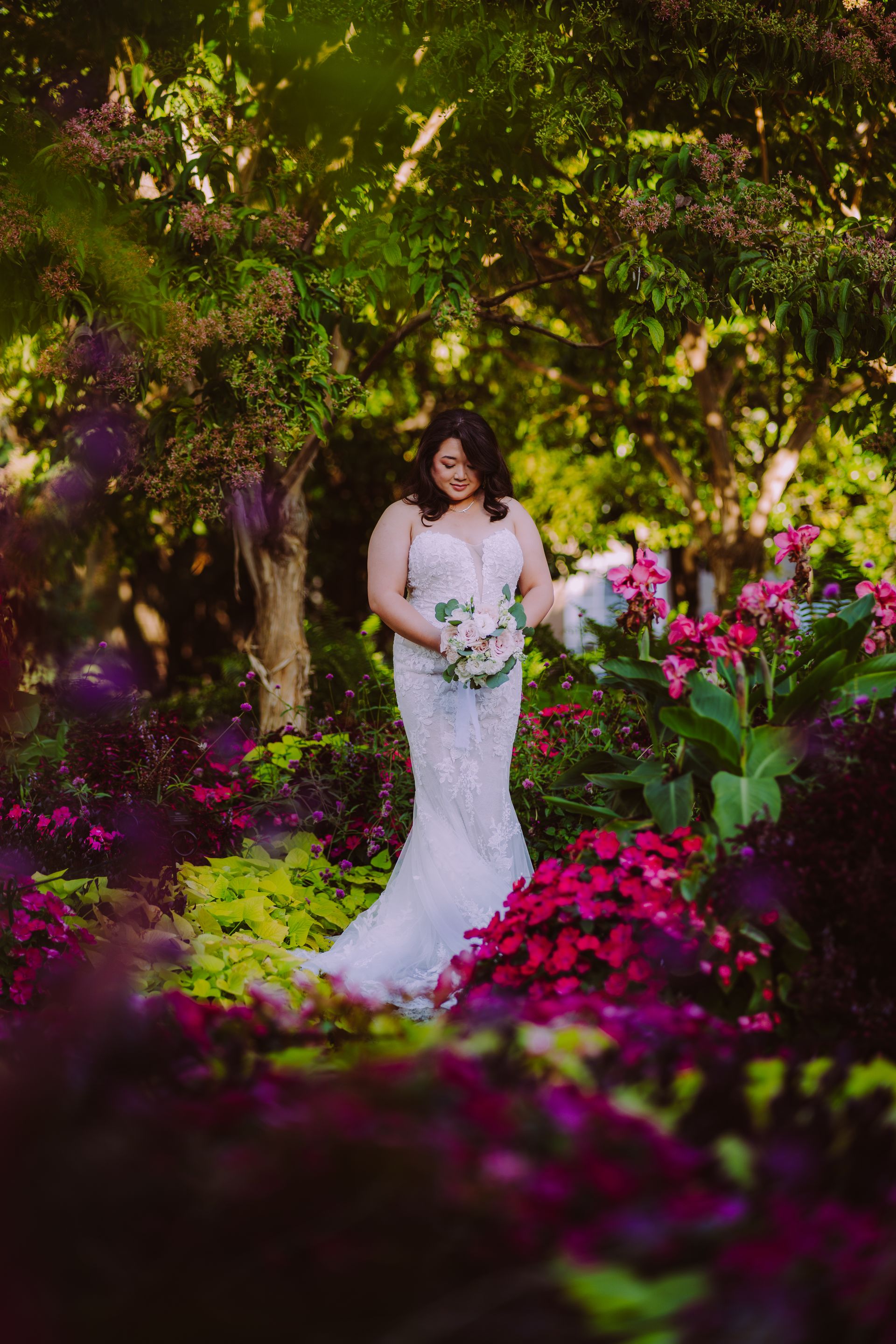 Bride in white gown holding flowers, standing amidst colorful garden, natural light.