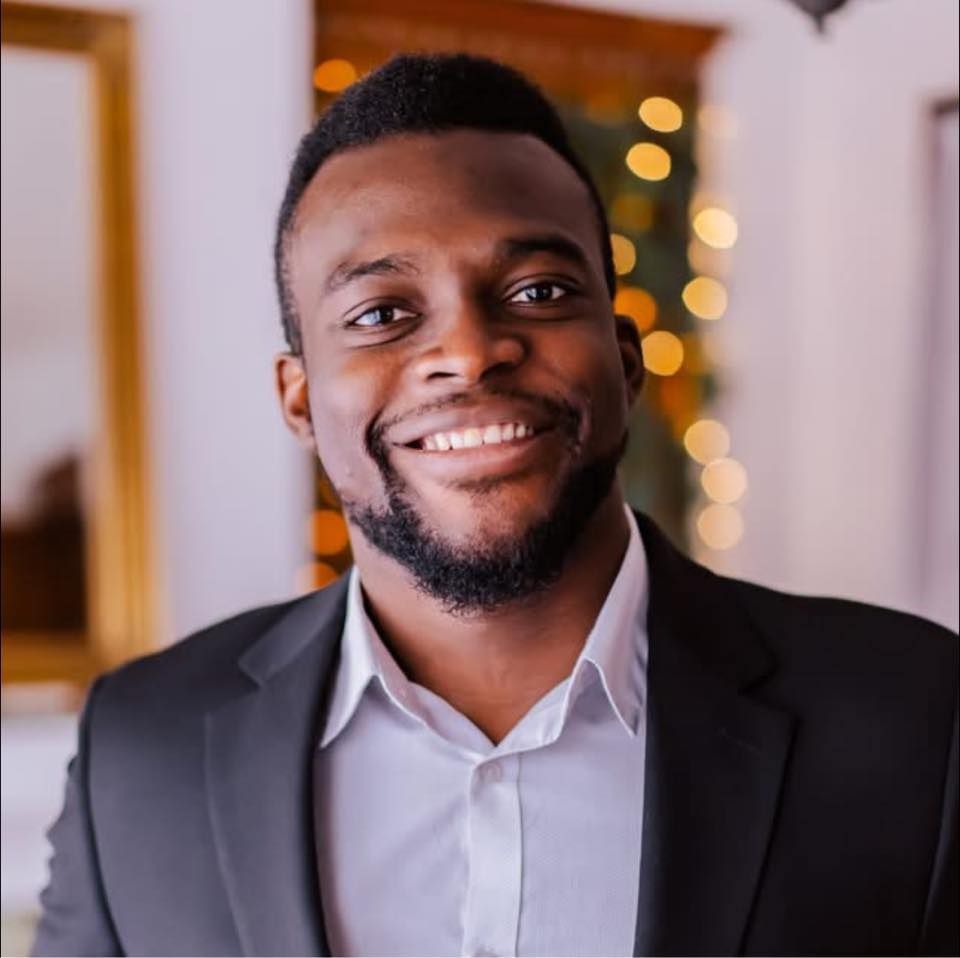 Man in black blazer and white shirt smiles at the camera, blurred background.