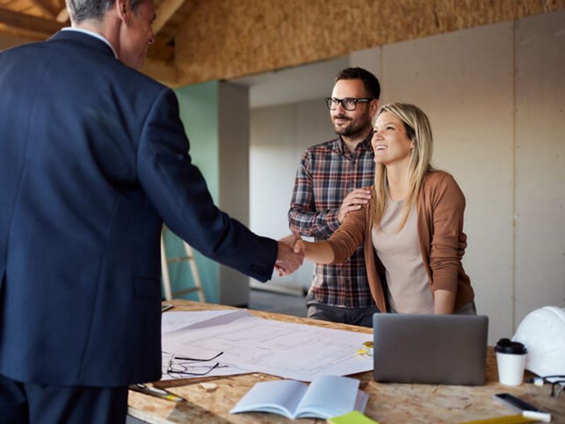 Business handshake across a desk with documents, laptop, and two people in an office meeting