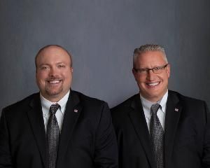 Two men in black suits and ties smiling against a gray studio backdrop