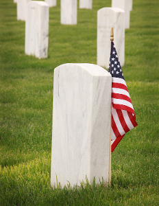 White gravestones in a cemetery, one marked with a small American flag on a green lawn