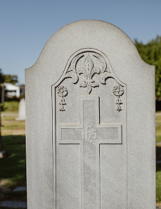 Gray gravestone with a carved cross and floral ornament, outdoors against a blue sky.