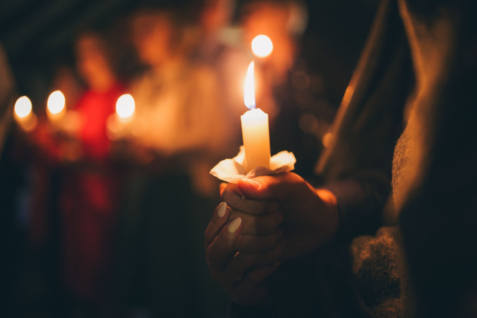Hands holding a lit candle at a dim vigil with blurred lights in the background