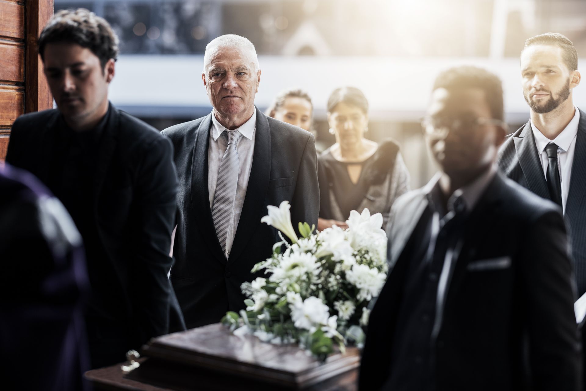 Mourners stand around a casket adorned with white flowers in a dim funeral chapel.