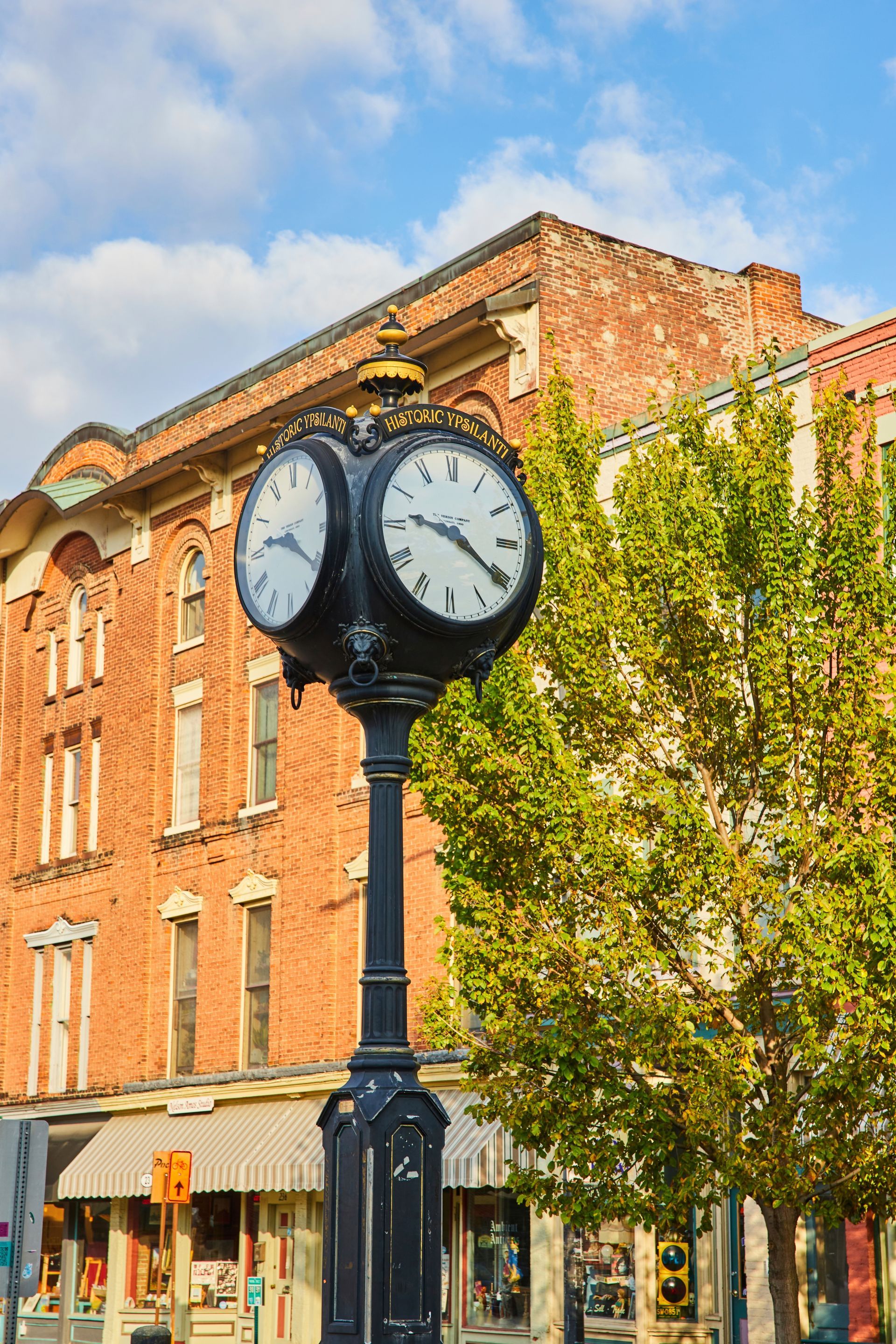 Black street clock in front of a red brick building and leafy tree under a blue sky