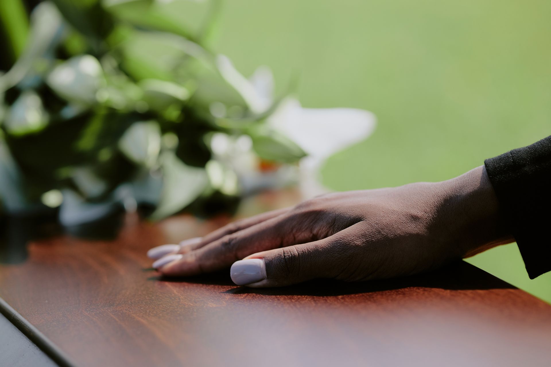 Hand resting on a wooden table beside a blurred green plant