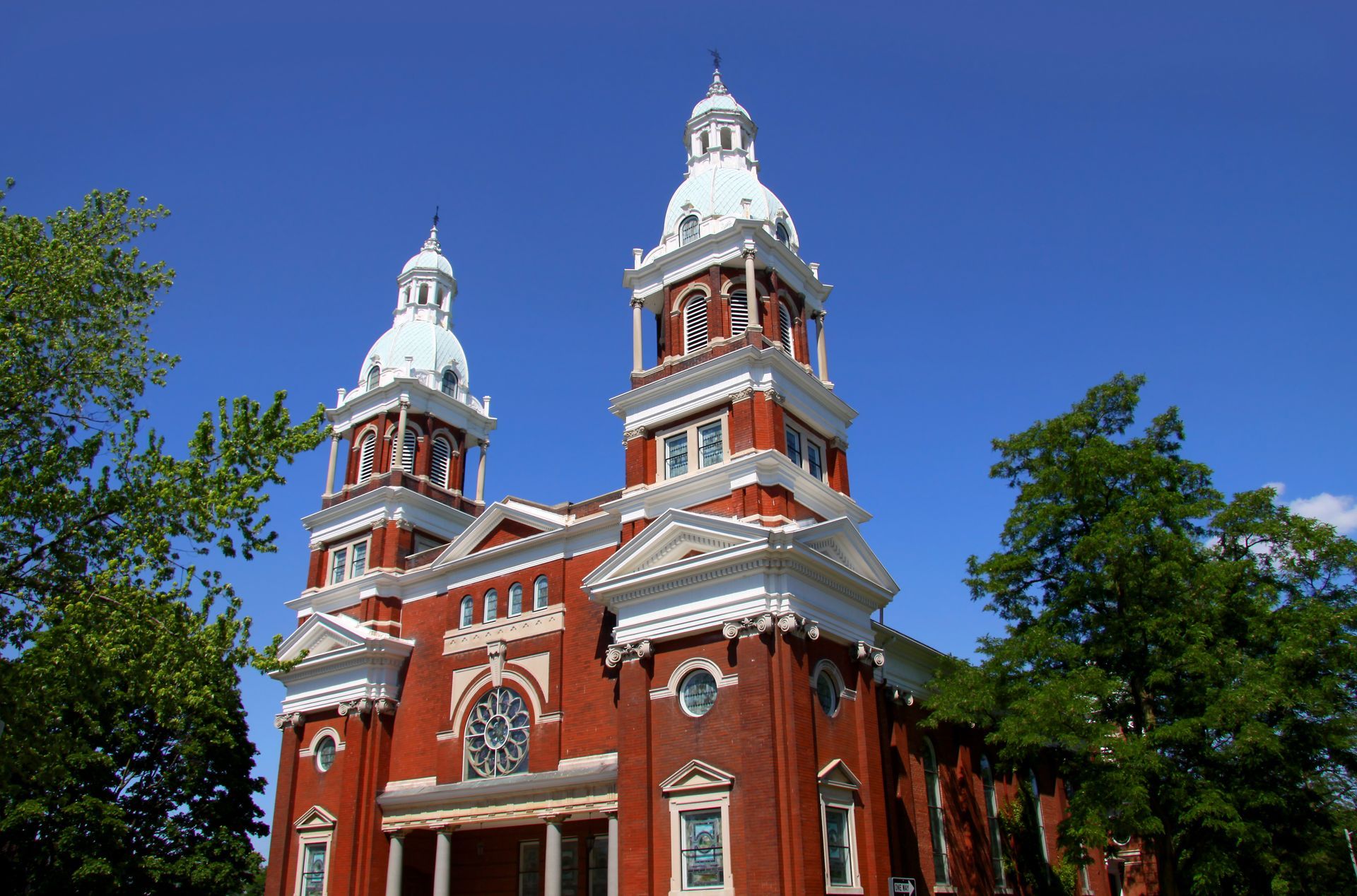Red brick church with twin white towers against a bright blue sky, framed by green trees