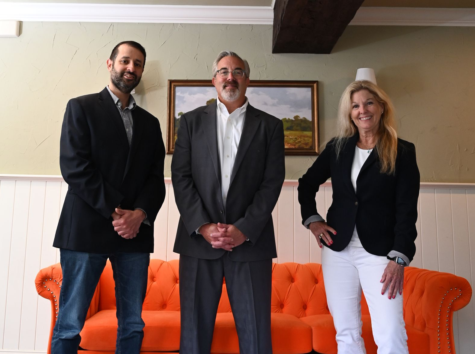 Three people standing indoors in suits in front of an orange couch, smiling at the camera.