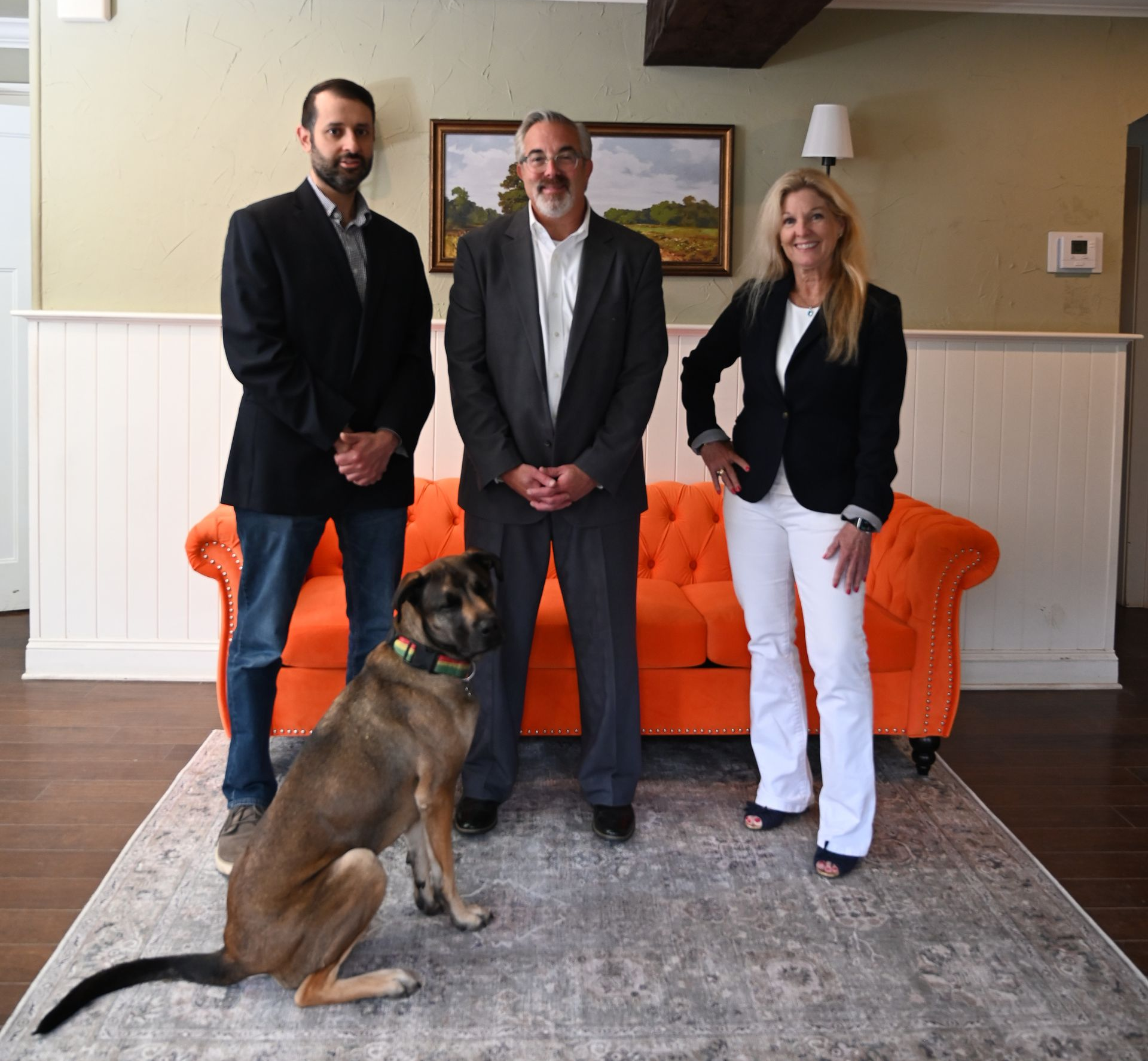Three people standing in a living room with an orange couch; a dog sits in the foreground.