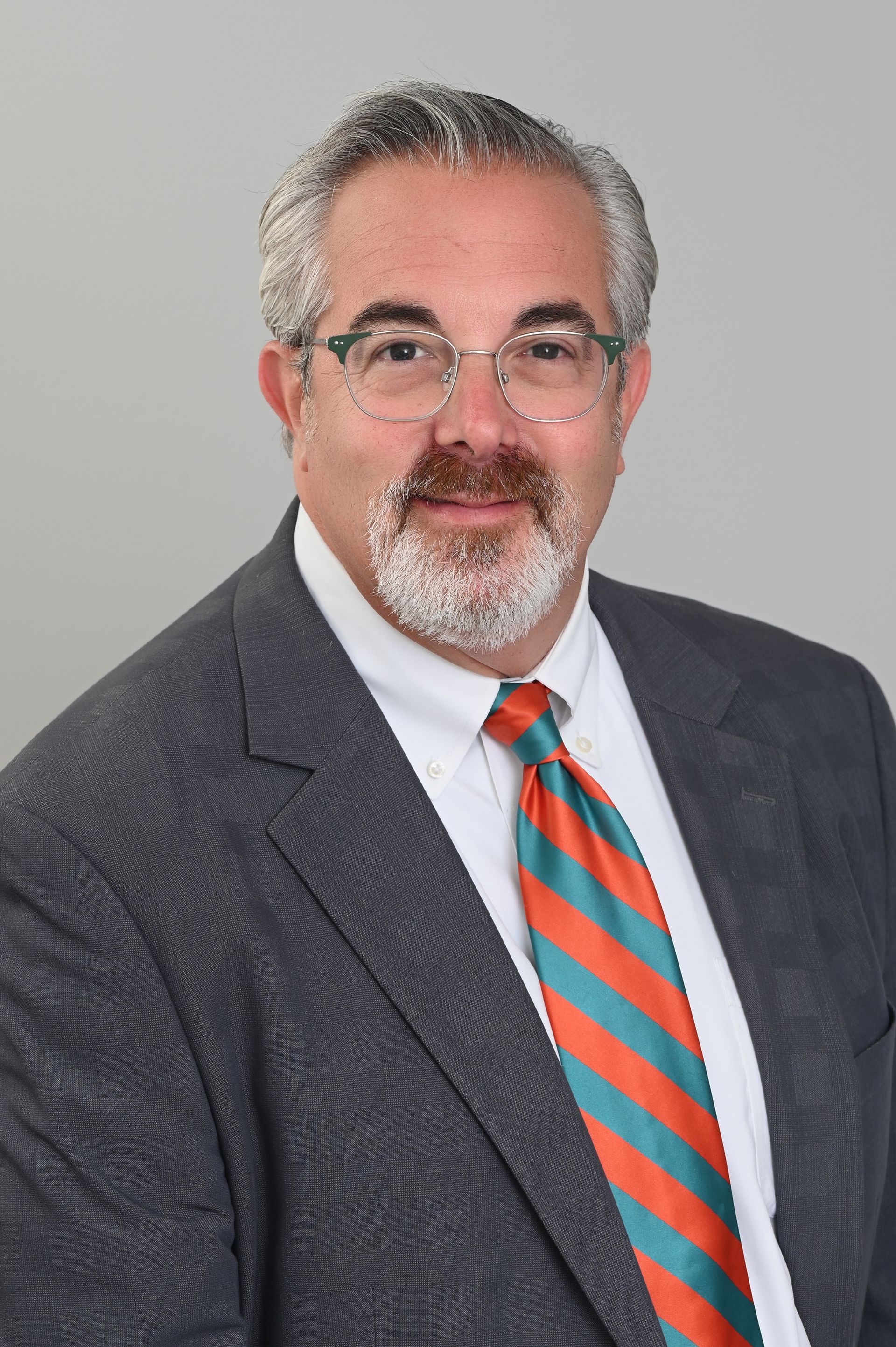 Studio portrait of a man in a dark suit, white shirt, and striped orange-blue tie against a gray background