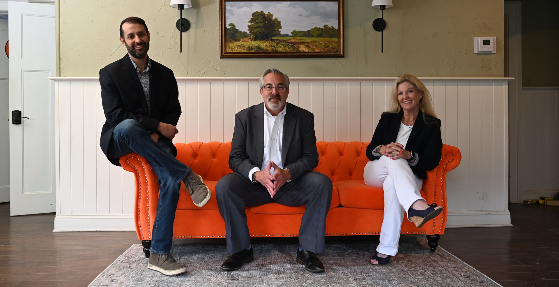 Three people seated in a room, with a bright orange couch and framed landscape painting behind them.