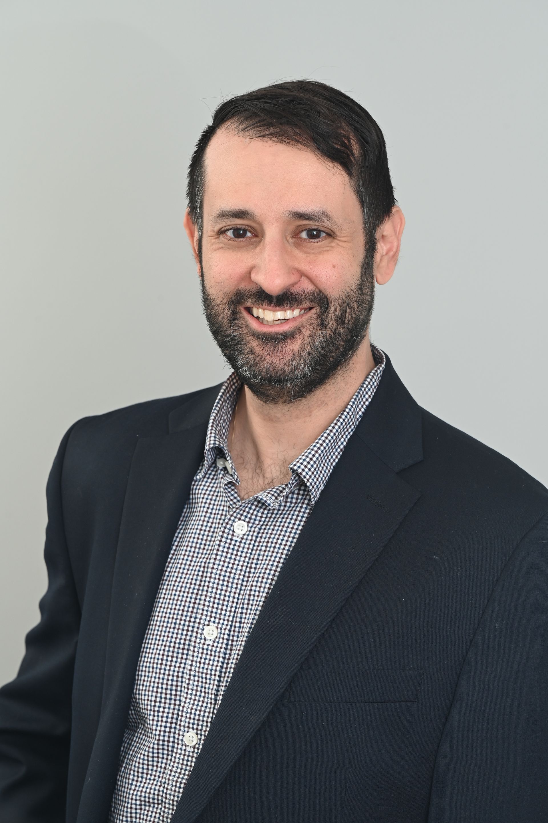 Smiling man in a dark blazer and patterned shirt against a light gray background