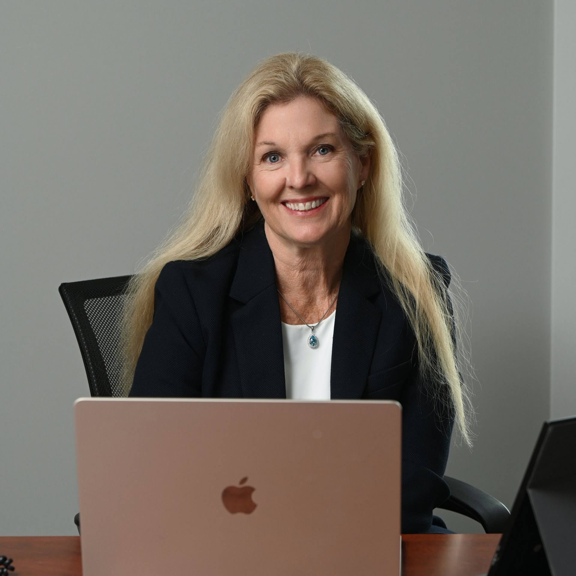 Smiling woman seated at a desk with a laptop in a plain office setting