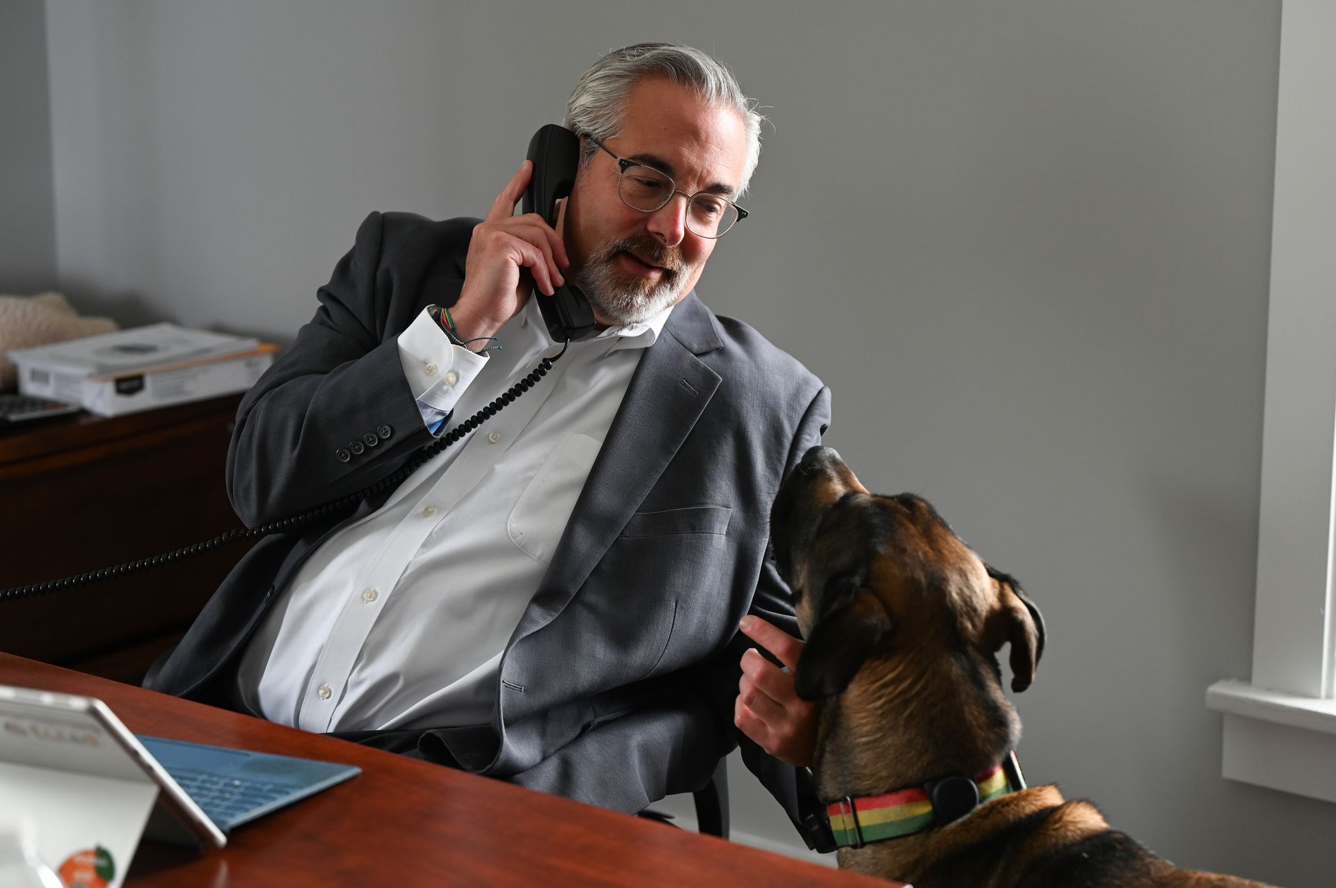 Man in a gray suit speaking on a phone at a desk while a dog looks up at him