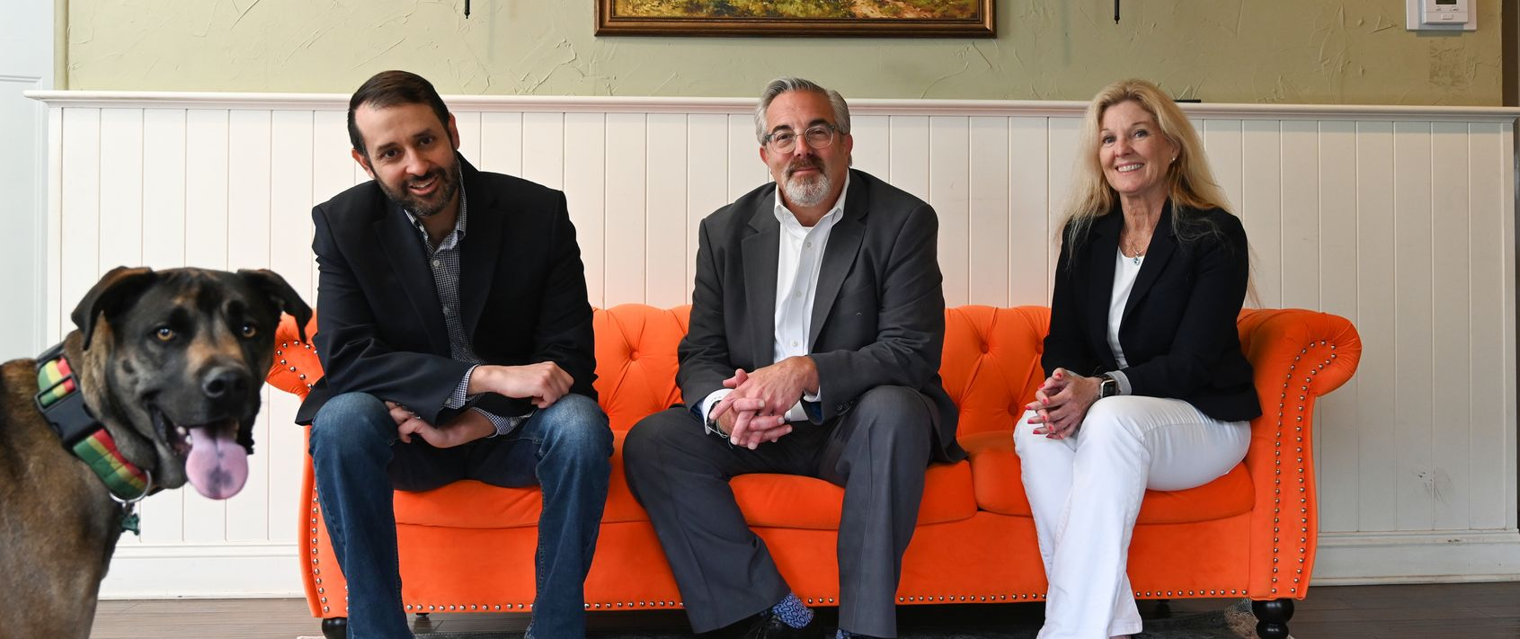 Three people sit on an orange couch with a dog in a living room, smiling at the camera.