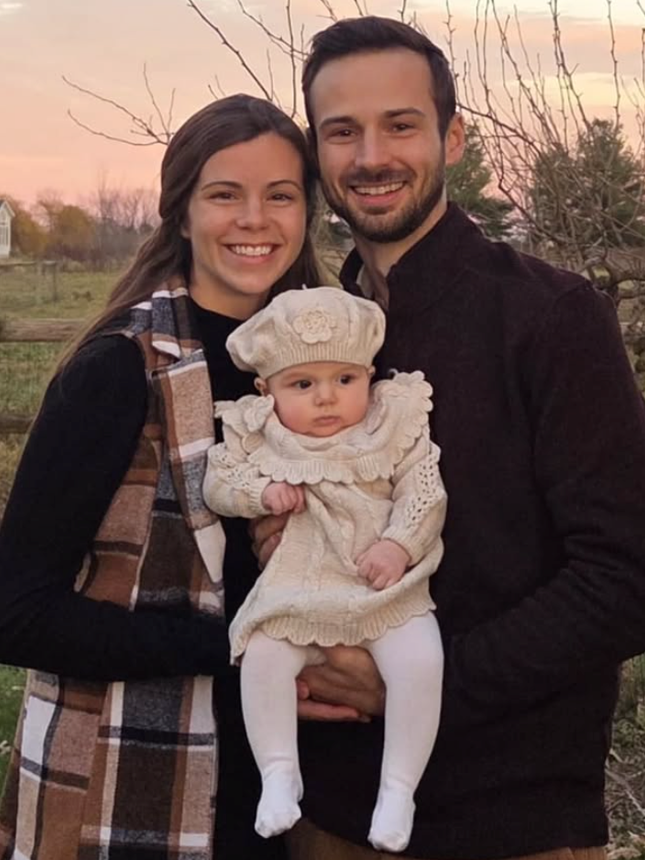 Smiling couple holding a baby outdoors at sunset, with a field and trees in the background.