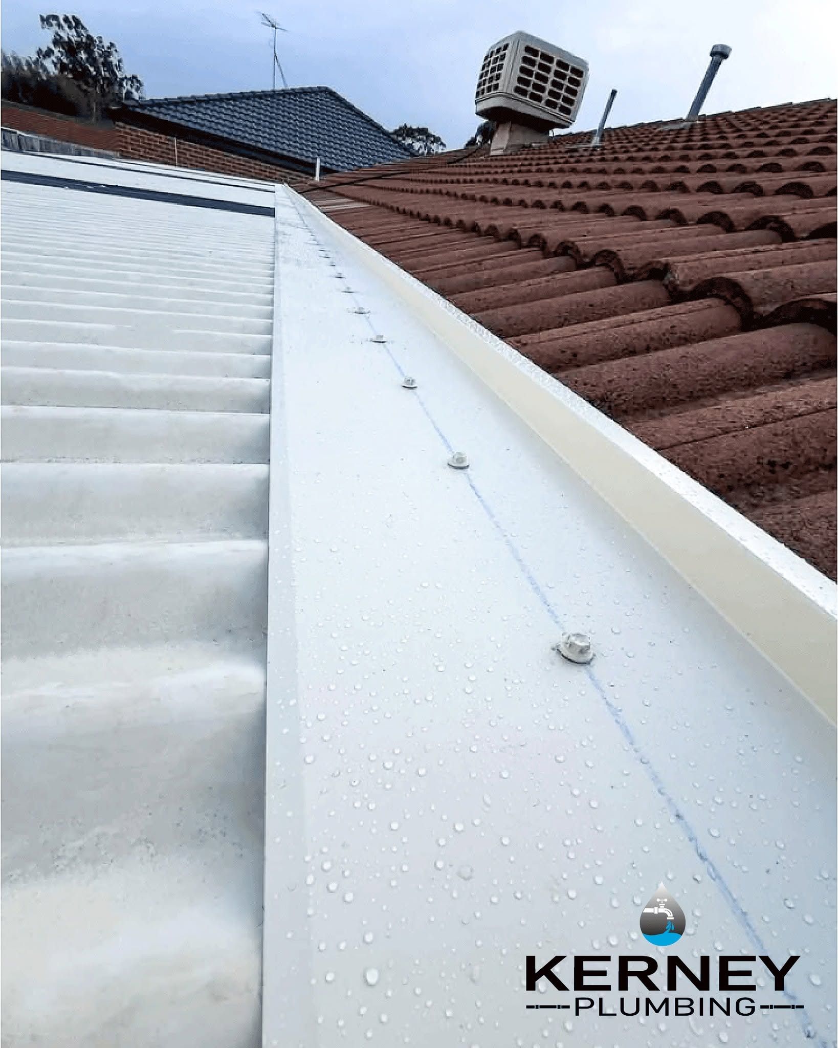 White Metal Roof Valley Between Dark Red Tiled Roofs Under Cloudy Sky — Kerney Plumbing in Bacchus Marsh, VIC