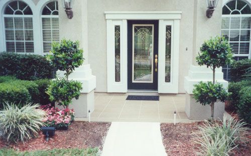 Exterior of a home with a front entrance. White pillars and door frame, black door, shrubs and trees in the yard.