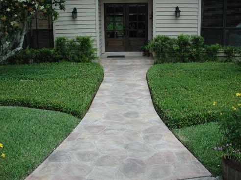 Stone walkway leading to double doors of a house, flanked by green lawns and shrubs.