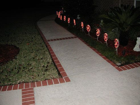 A brick-lined sidewalk curves, lit by red lights. Lawn on the left, bushes and dark space on the right.