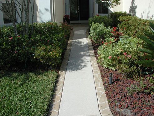 Concrete walkway leading to a front door, flanked by bushes and landscaping.