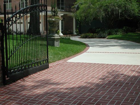Black gate opens to brick driveway and grassy lawn, leading to a house with a porch.