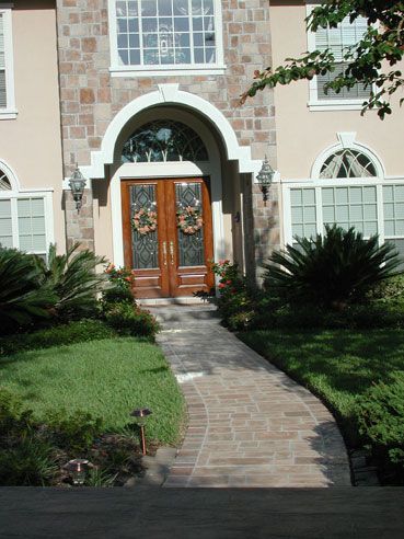 Pathway to a two-door entrance, flanked by landscaping. Beige stucco home with arched doorway.