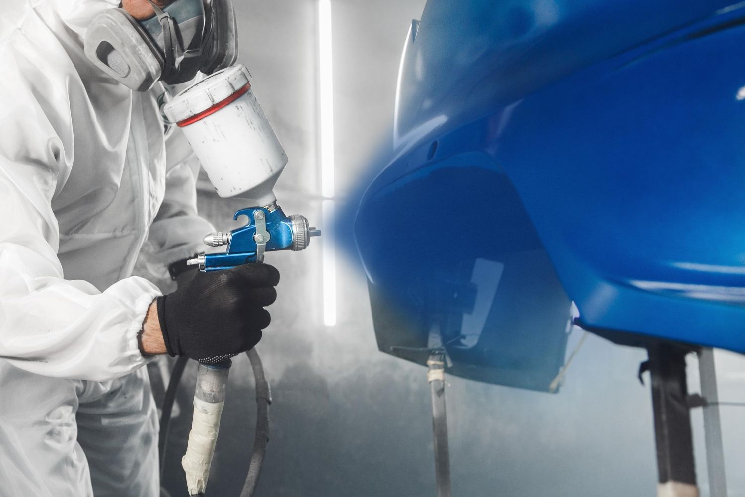 Person in protective suit spraying blue paint onto a car part in a spray booth.