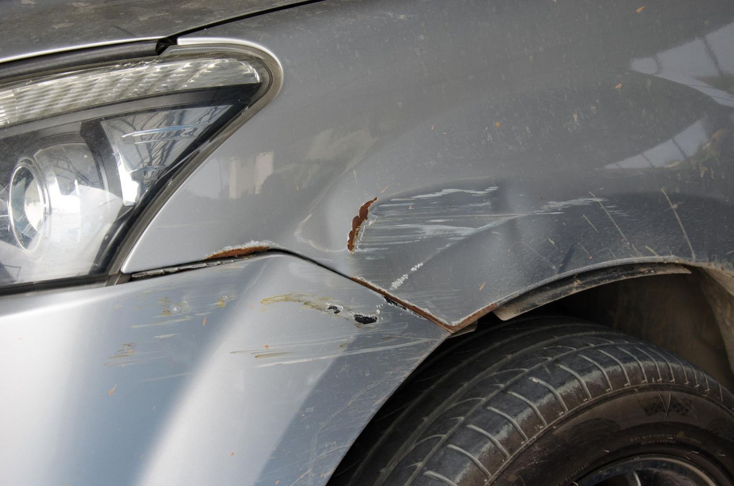 Close-up of a grey car with paint scratches and rust along the seam between the front bumper and fender.