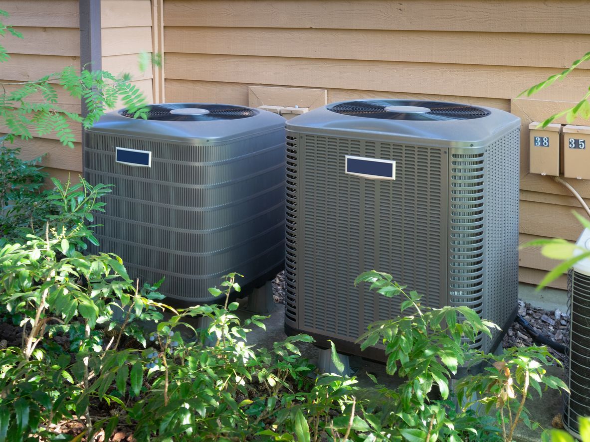 Two air conditioners are sitting next to each other on the side of a house.