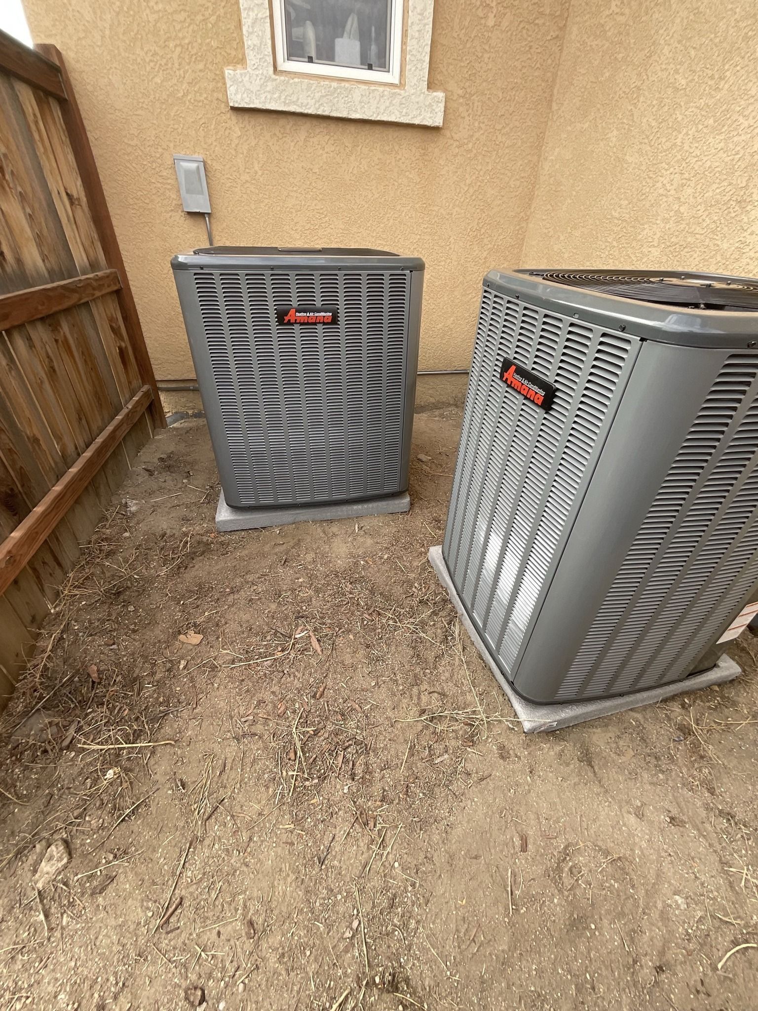 Two air conditioners are sitting in the dirt in front of a house.