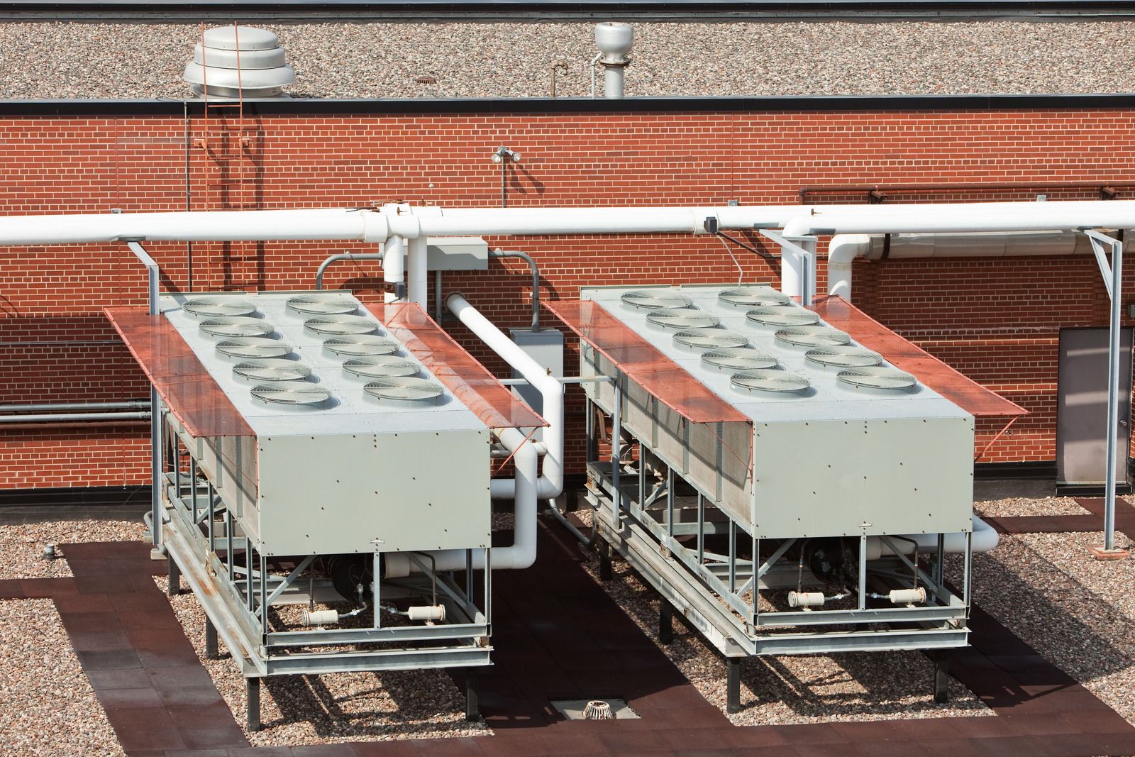 Two rooftop air conditioning units with fans, connected by white pipes, against a brick background.