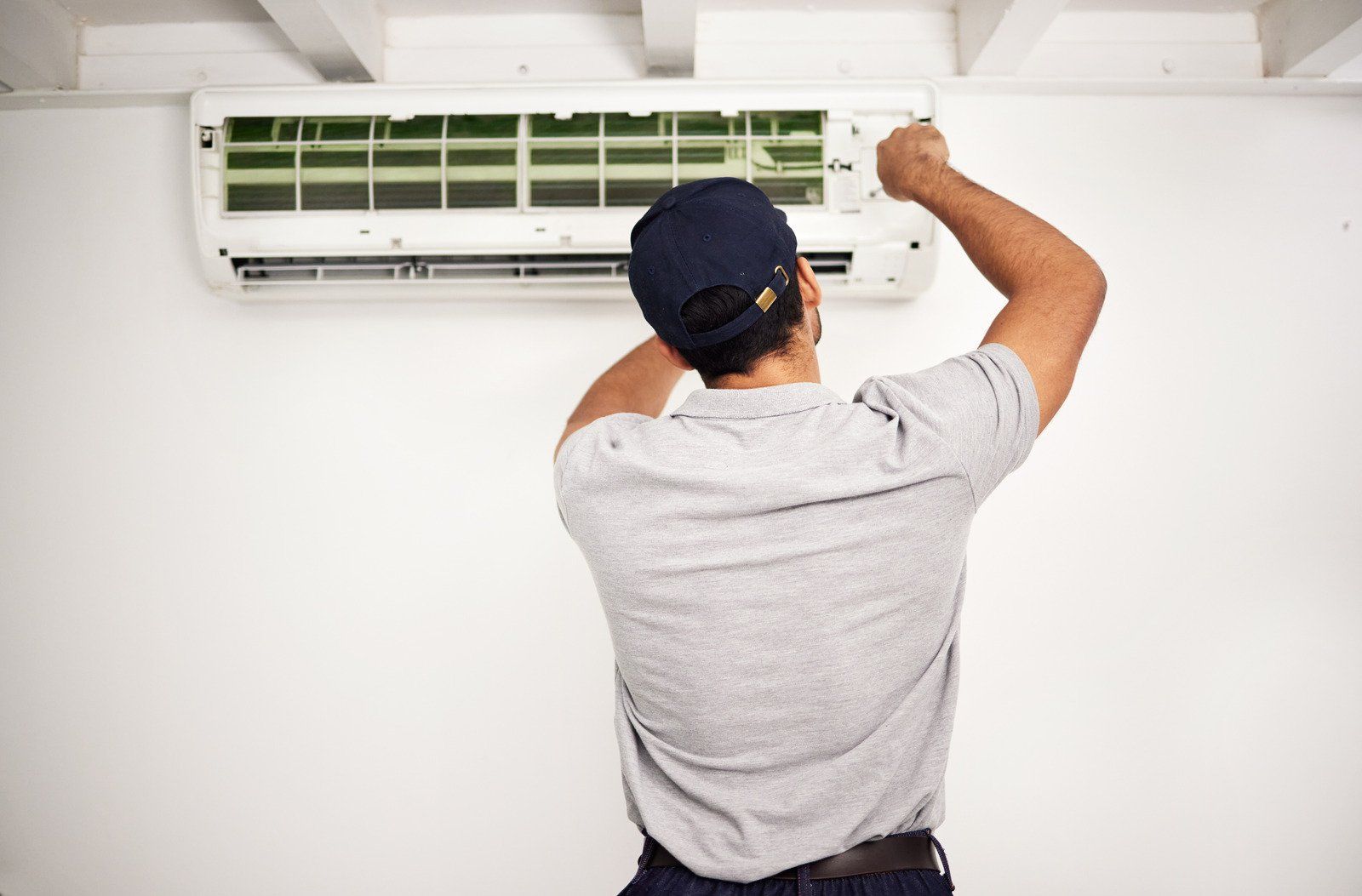 Person in cap repairing air conditioner on a white wall.