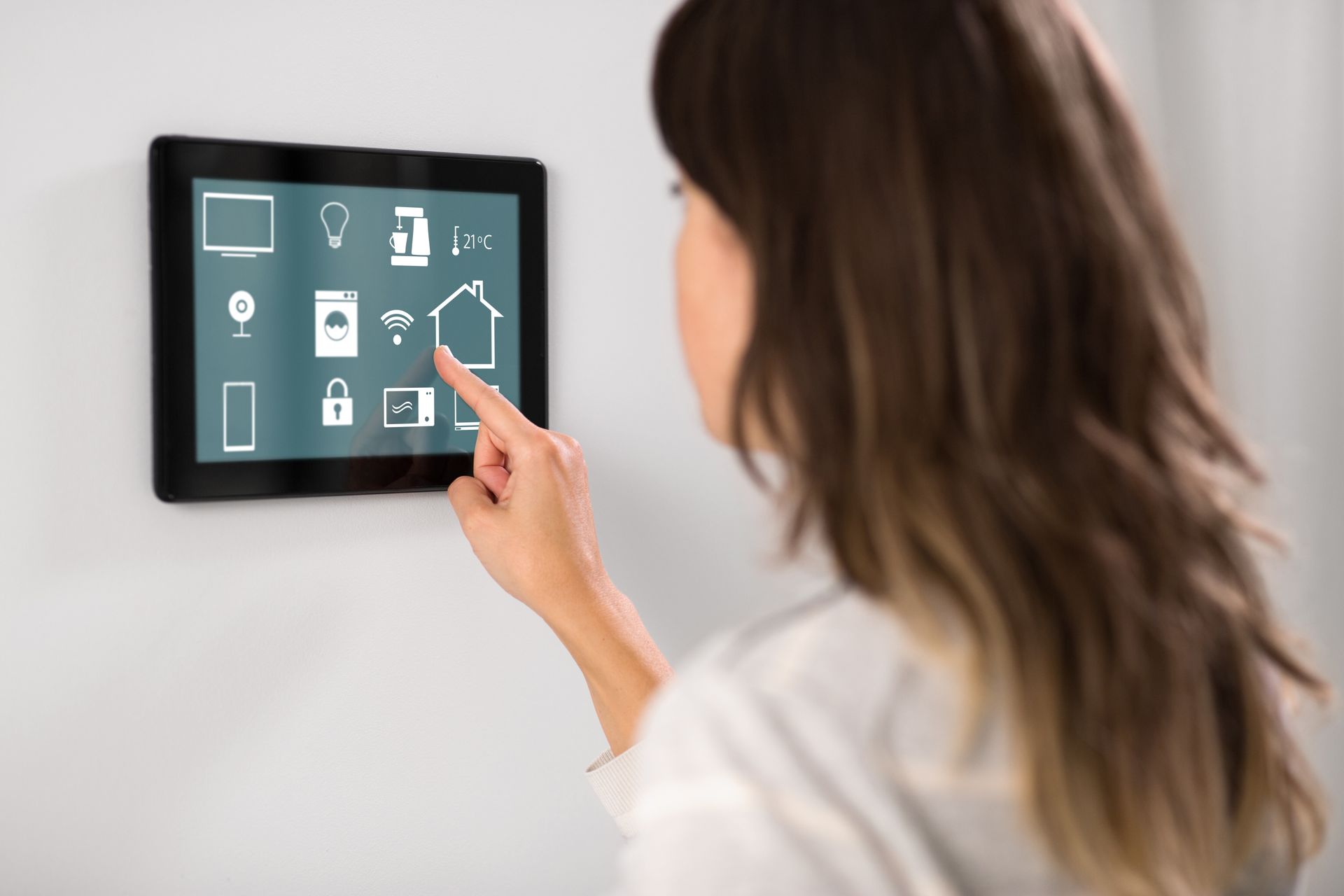 Een persoon tikt op een aan de muur gemonteerd bedieningspaneel voor een slim huis met verlichte pictogrammen.