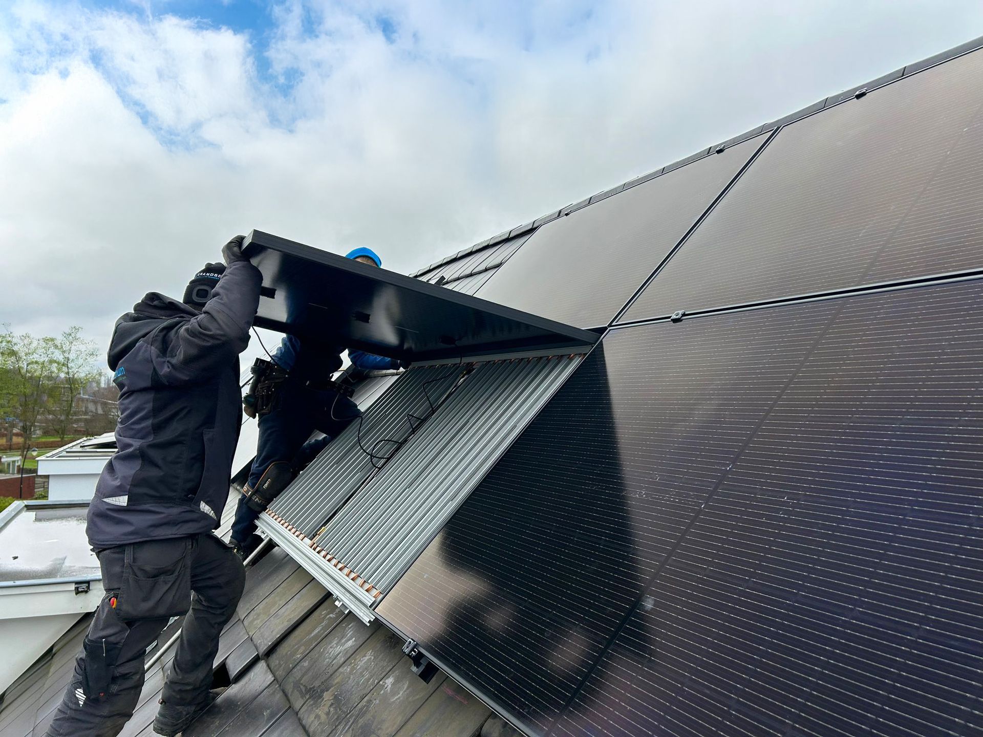 Arbeiders openen een ventilatieopening op het dak van een donker metalen gebouw onder een bewolkte hemel.